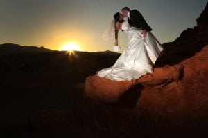 A bride and groom kissing on a rock at sunset.
