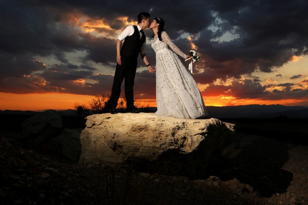 A bride and groom standing on a rock at sunset.