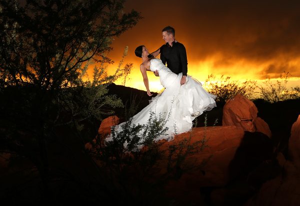 A bride and groom standing on a rock at sunset.