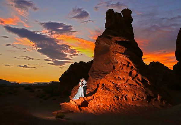 A bride and groom standing in front of a rock formation at sunset.
