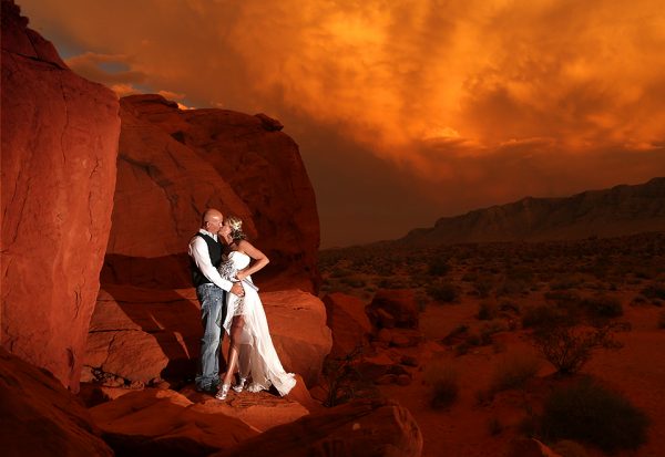 A bride and groom pose in front of a red sunset in the desert.