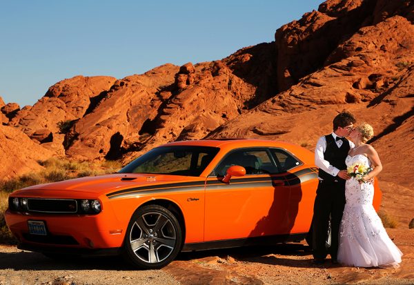 A bride and groom standing next to an orange car.