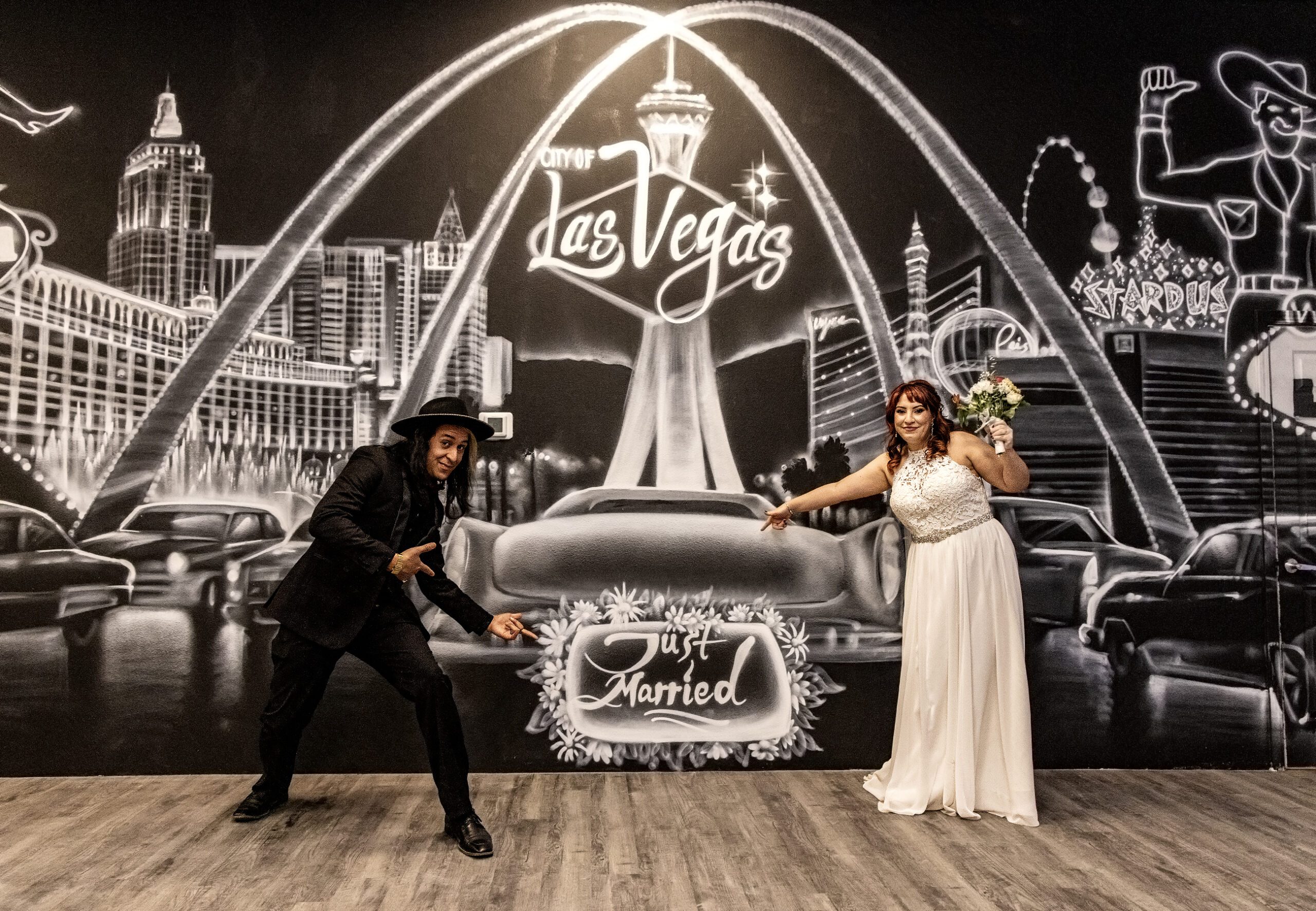 A newlywed couple celebrates in front of a Vegas chapel-themed mural, striking playful poses with the bride holding a bouquet.